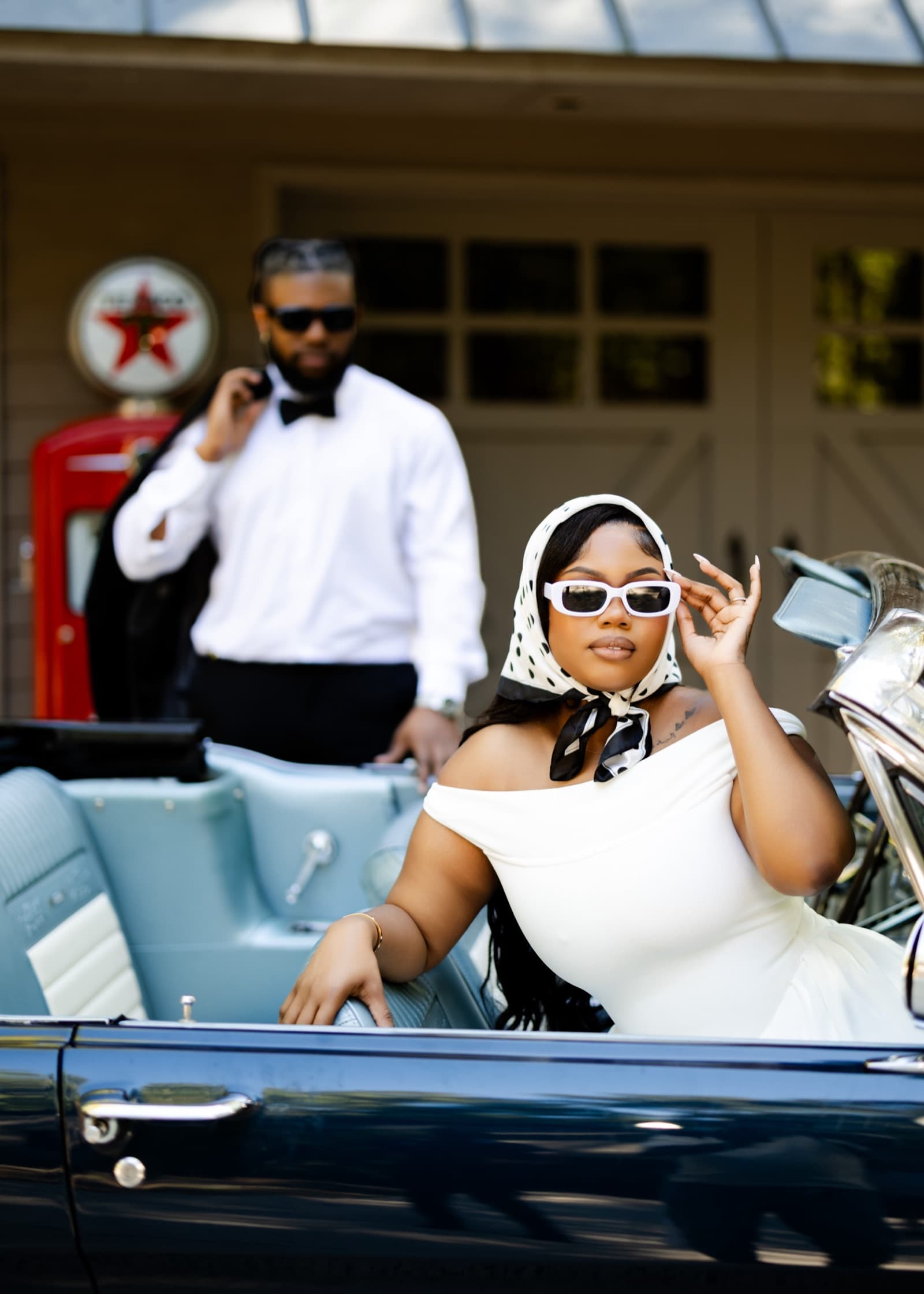 Kijori standing by a vintage convertible in black tie while Amoy sits in the car in a headscarf and sunglasses